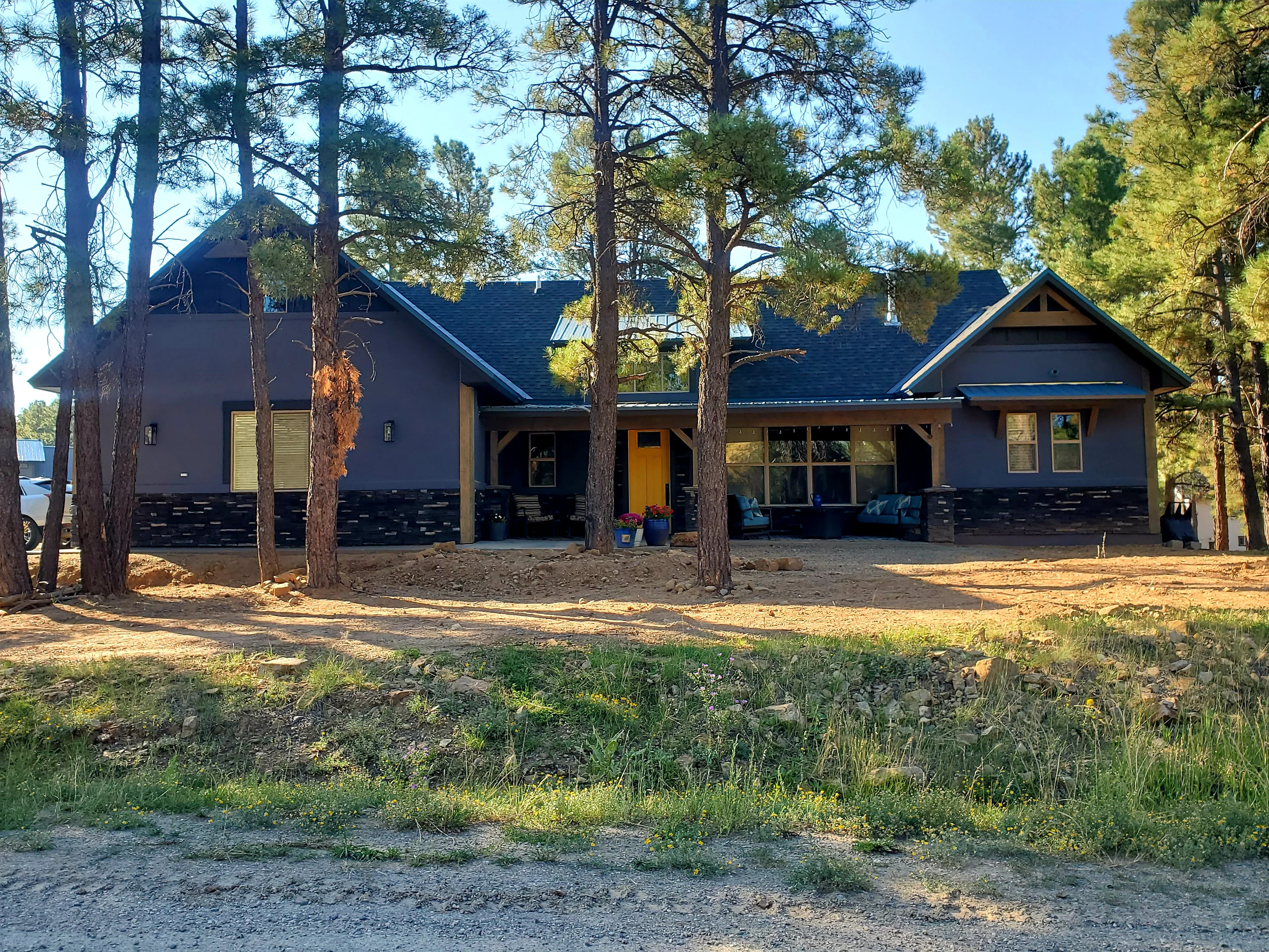 Dark stucco home with stone foundation, asphalt shingle roof, and covered porches among pine trees