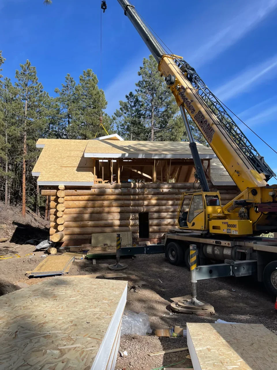 Log cabin under construction with yellow crane lifting roof frame, timber walls, and foundation work visible