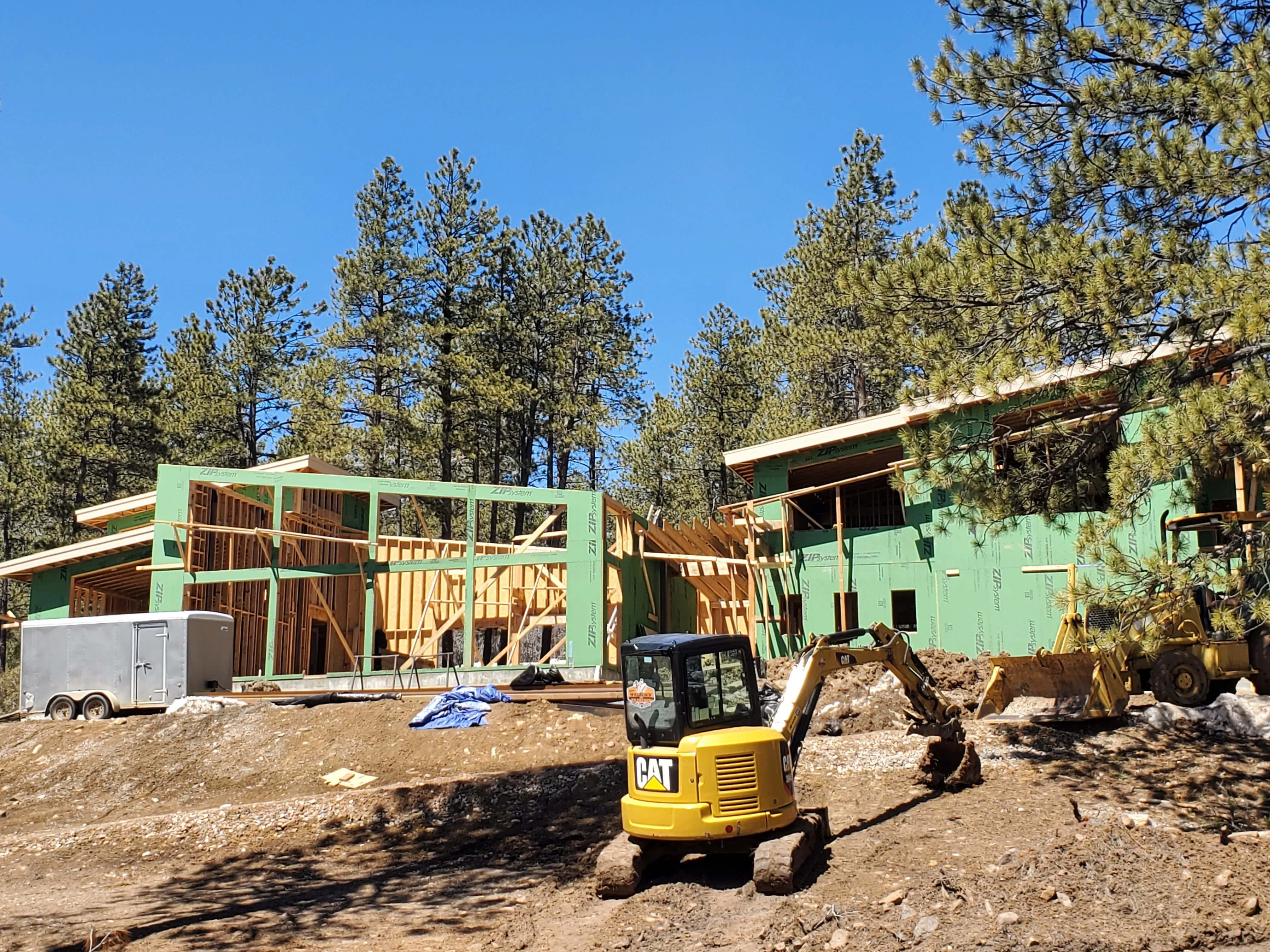 Wood-framed residential structure with green house wrap, exposed trusses, and active site excavation
